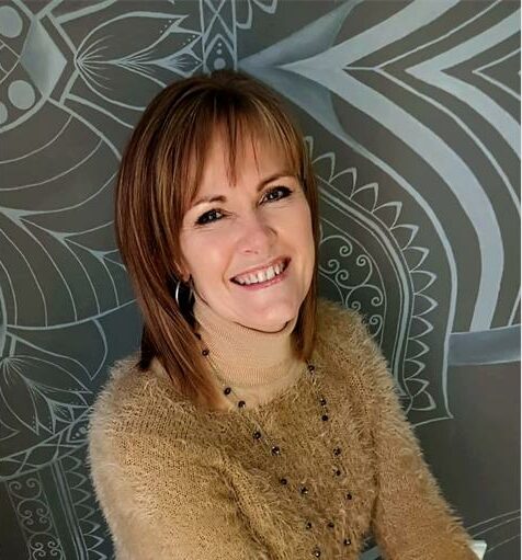A white woman with ginger hair wearing a camel coloured sweater and necklace smiling at the camera with a grey and white patterned background.