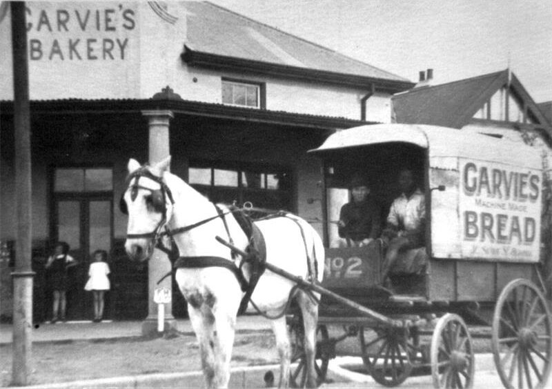 A black and white photo of a building and horse-drawn cart.