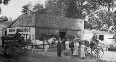 A black and white photo of an old bakery building, horse-drawn cart and people standing in front of the building.
