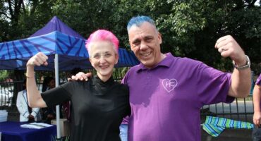 Woman wearing black with pink hair flexes her arm along with a man wearing a purple shirt and blue hair