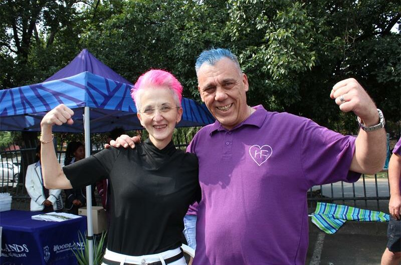 Woman wearing black with pink hair flexes her arm along with a man wearing a purple shirt and blue hair