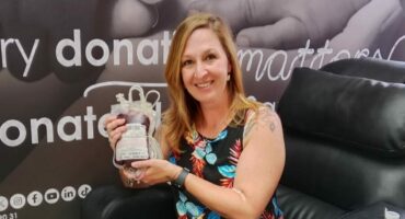 A white woman dressed in black pants and a floral shirt sitting on a black leather blood donation chair holding a gift hamper.