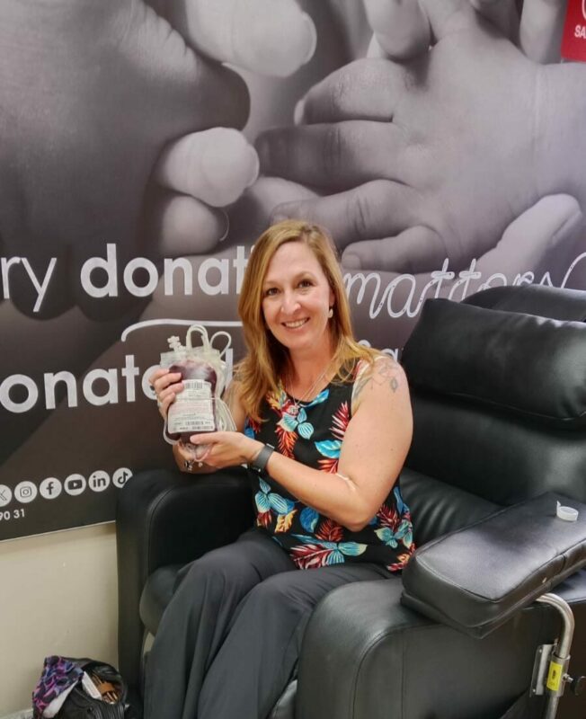 A white woman dressed in black pants and a floral shirt sitting on a black leather blood donation chair holding a gift hamper.