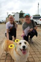 Two women standing with a dog wearing sunflowers