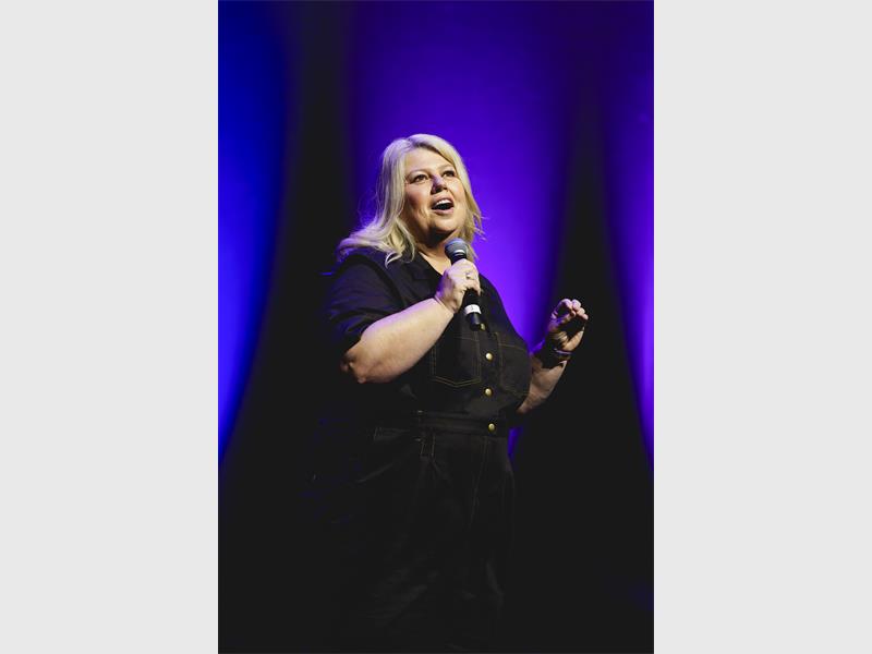 A woman with blonde hair on stage with a microphone in her hand, all dressed in black. 