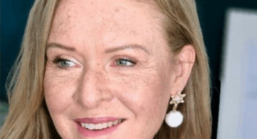 A woman with light brown hair and freckles smiles warmly. She wears star-shaped earrings with white pendants and a red top, conveying a cheerful tone.