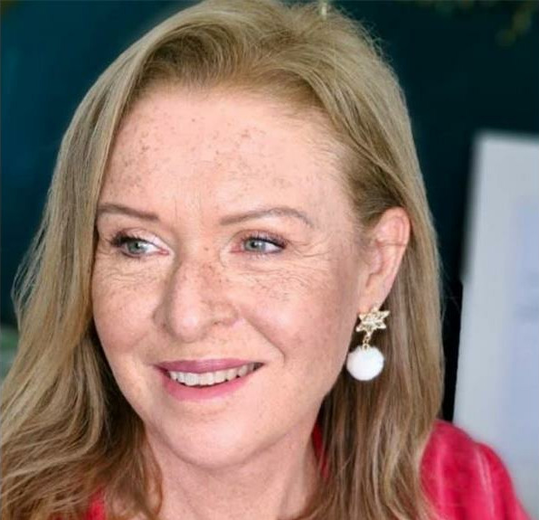 A woman with light brown hair and freckles smiles warmly. She wears star-shaped earrings with white pendants and a red top, conveying a cheerful tone.