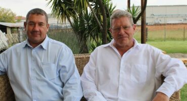 Two white men sitting on a couch in a garden setting