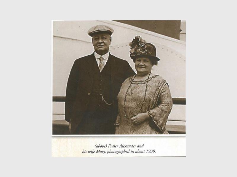 A black and white image of a couple on the deck of a ship, dressed in a suit and elegant dress and hats.