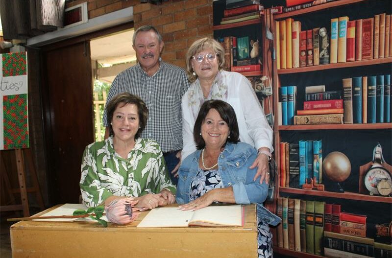 Vier wit mense, een man en drie vrouens sit by 'n hout skoolbank