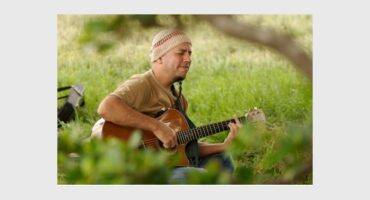A man wearing a knitted beanie and beige T-shirt playing the guitar in a tranquil greenery setting.