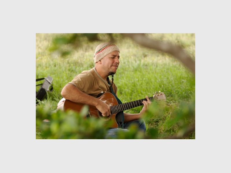 A man wearing a knitted beanie and beige T-shirt playing the guitar in a tranquil greenery setting.