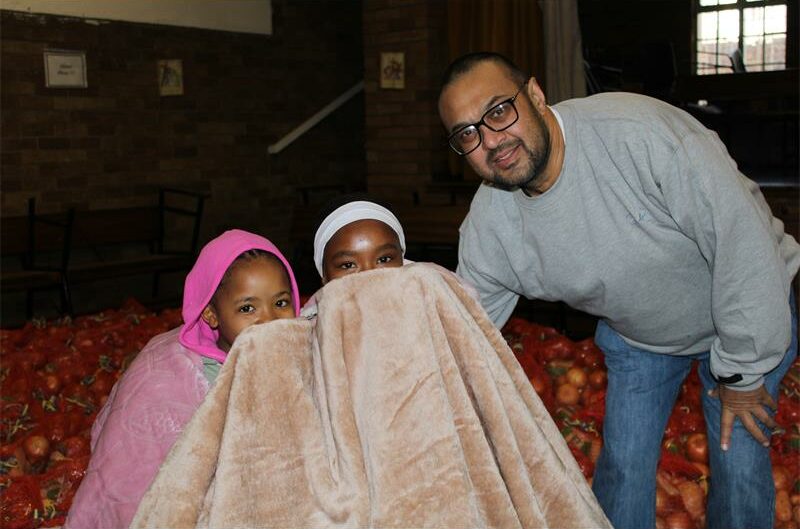Indian man wearing glasses, stands next to two girls wrapped in an blanket with their faces covered