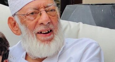 A Muslim man dressed in traditional clothing with a grew beard and wearing glasses.