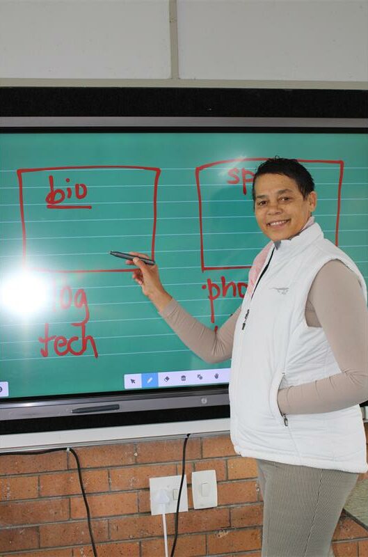 Coloured woman with short hair stands in front of a digital school board