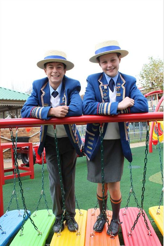 White boy and white girl wearing school uniforms and straw hats lean over a red bar of playground equipment