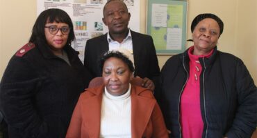 Three black women and a black male posing for a photograph, with one woman seated in front of the three others standing.
