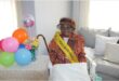 A granny wearing a happy birthday sash sitting next to a table with balloons