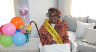 A granny wearing a happy birthday sash sitting next to a table with balloons