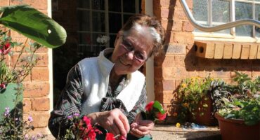 Bejaarde vrou sit op die stoep tussen blomme