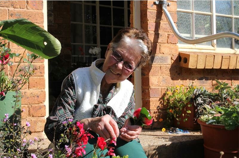 Bejaarde vrou sit op die stoep tussen blomme