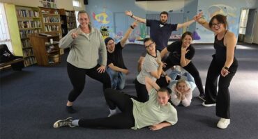A troupe of actors rehearsing for a show in various poses, mostly wearing black pants and grey shirts.
