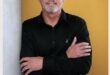 A white middle-aged man wearing all black clothing standing in front of a yellow background smiling.
