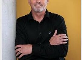A white middle-aged man wearing all black clothing standing in front of a yellow background smiling.