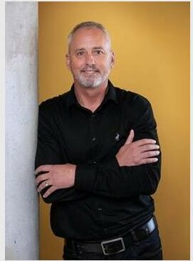 A white middle-aged man wearing all black clothing standing in front of a yellow background smiling.