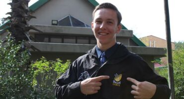 Young man stands in front of building pointing to the emblem on his jacket