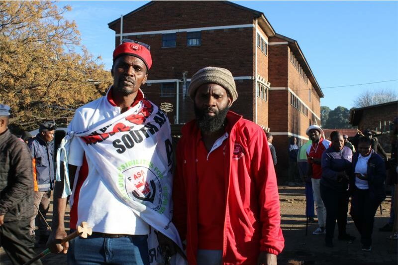 Two black men stand side-by-side with a brick building in the background