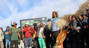 Black man stands in front of a group of protesters wearing traditional Zulu attire