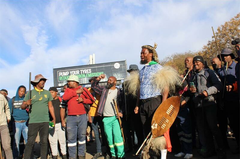 Black man stands in front of a group of protesters wearing traditional Zulu attire