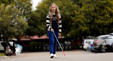 Woman with blonde hair walks through a parking area holding a white cane