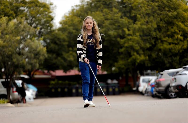 Woman with blonde hair walks through a parking area holding a white cane