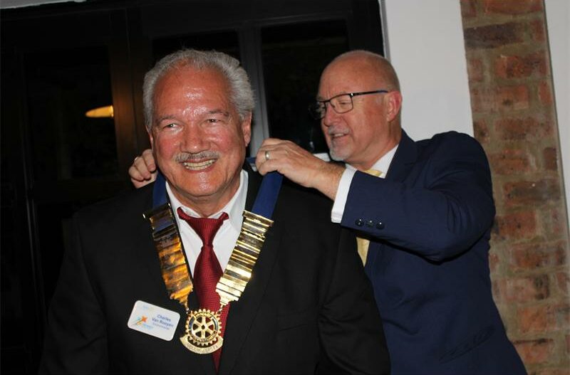 Grey haired man smiles as a man wearing glasses puts a chain around his neck