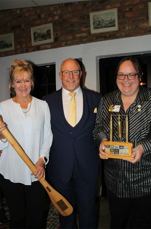 Man with glasses stand with a woman holding a large wooden spoon and another holding a trophy