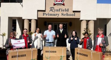 Group of adults and learners in school uniform standing behind boxes in front of a school