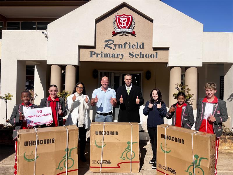 Group of adults and learners in school uniform standing behind boxes in front of a school