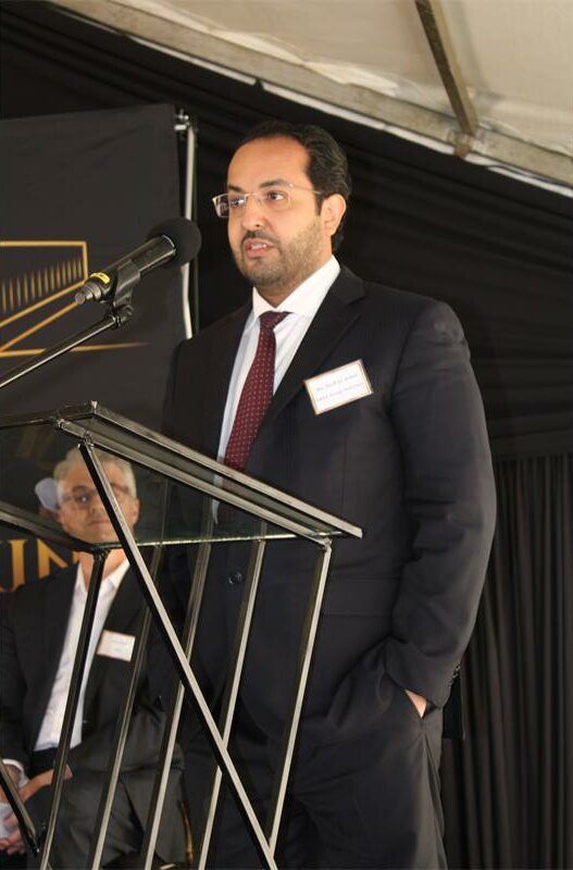 Arabic man wearing a black sui standing with his hands in his pockets while speaking into a microphone