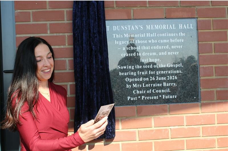 Woman with long dark hair, wearing a red dress reads from a paper next to a plaque