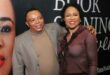 Black man wearing a suit has his hand draped over a black woman's shoulders