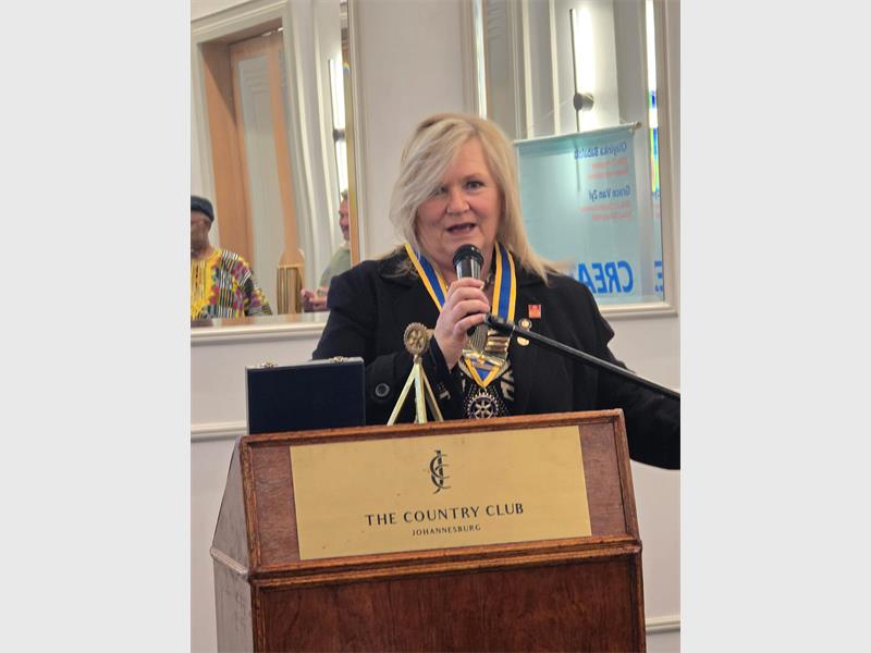 A white woman with blonde hair speaking at a podium while holding a microphone.