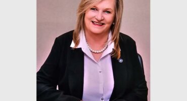 A blonde white woman wearing a black jacket and light shirt.