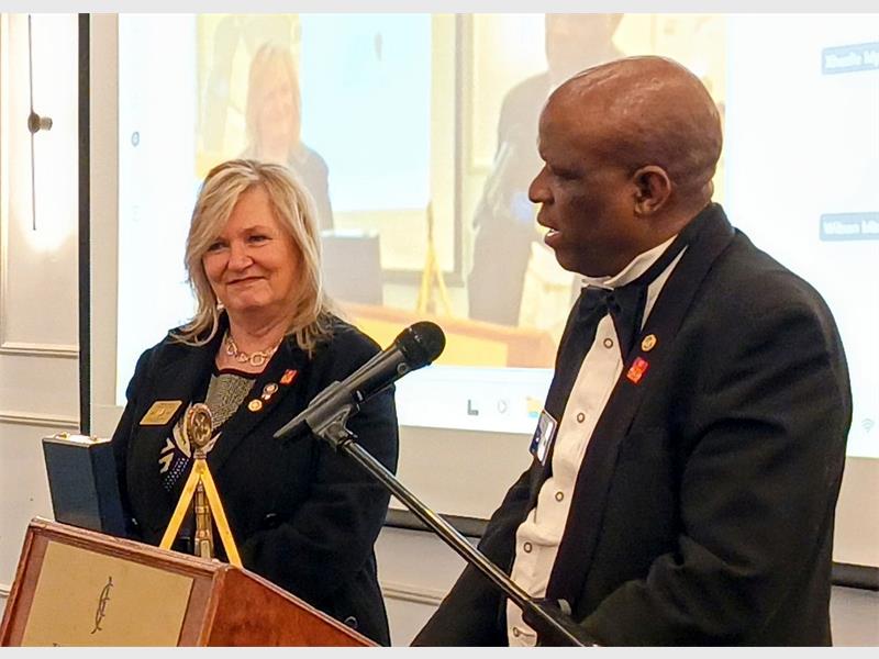A white woman next to a podium with microphone where a black man is speaking to her.