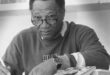 A black and white image of a black man wearing glasses, sitting at a desk with his arm resting on the desk next to a book.