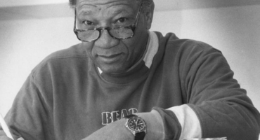 A black and white image of a black man wearing glasses, sitting at a desk with his arm resting on the desk next to a book.