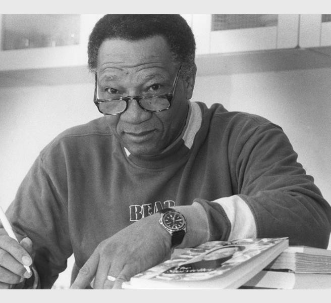 A black and white image of a black man wearing glasses, sitting at a desk with his arm resting on the desk next to a book.