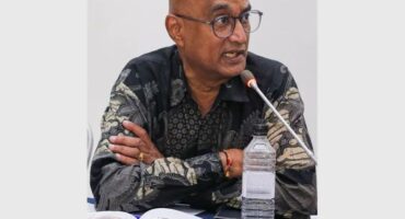 An Indian male sitting behind a desk with a bottle of water and papers on it, with his arms crossed and speaking into a microphone.