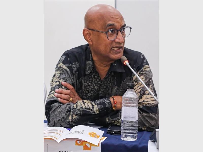 An Indian male sitting behind a desk with a bottle of water and papers on it, with his arms crossed and speaking into a microphone.